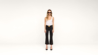 A woman wearing sunglasses, a white tank top, black pants, and black heels stands against a plain white background.