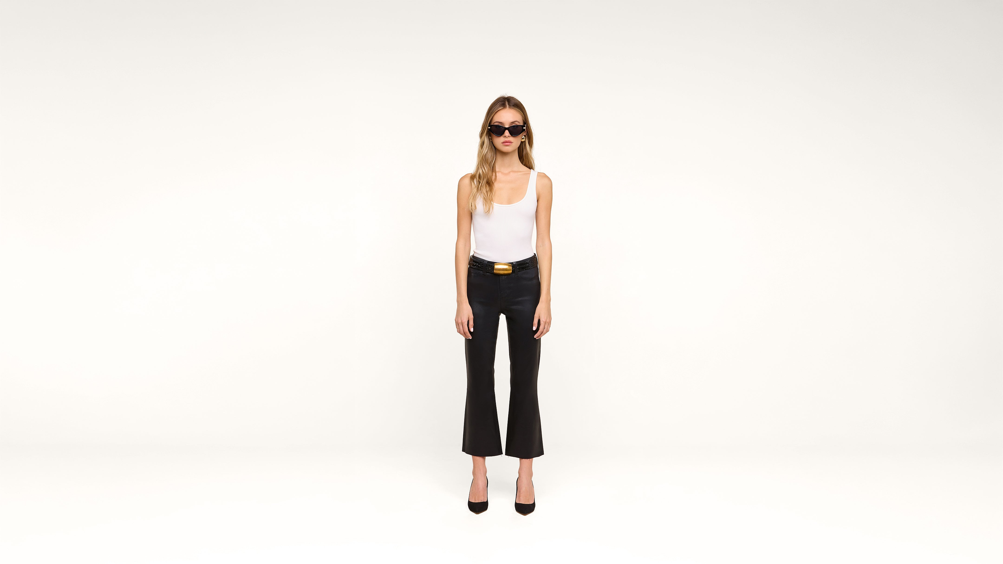 A woman wearing sunglasses, a white tank top, black pants, and black heels stands against a plain white background.