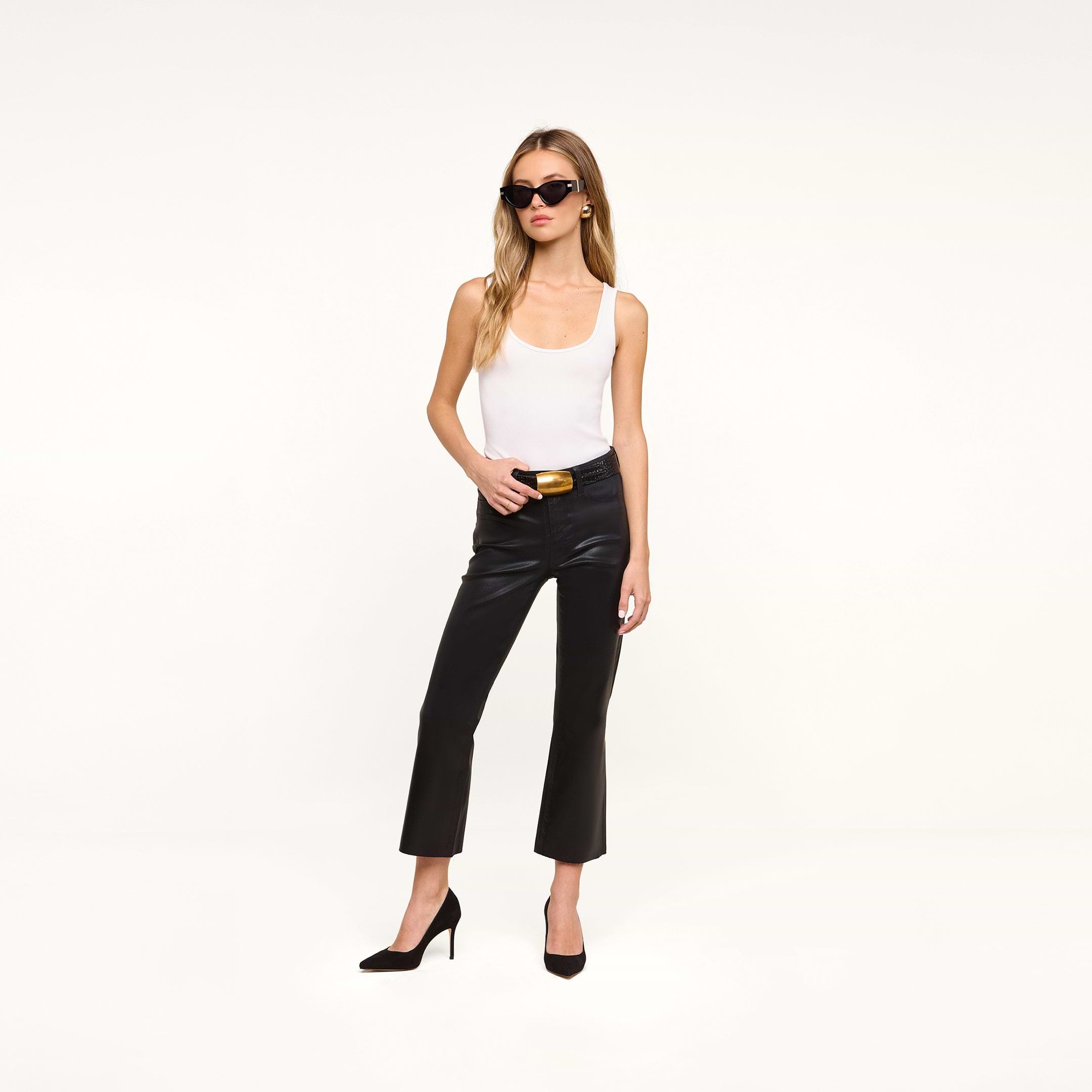 A woman wearing sunglasses, a white tank top, black Petite Kendra Coated Jeans, and black heels stands against a plain white background.