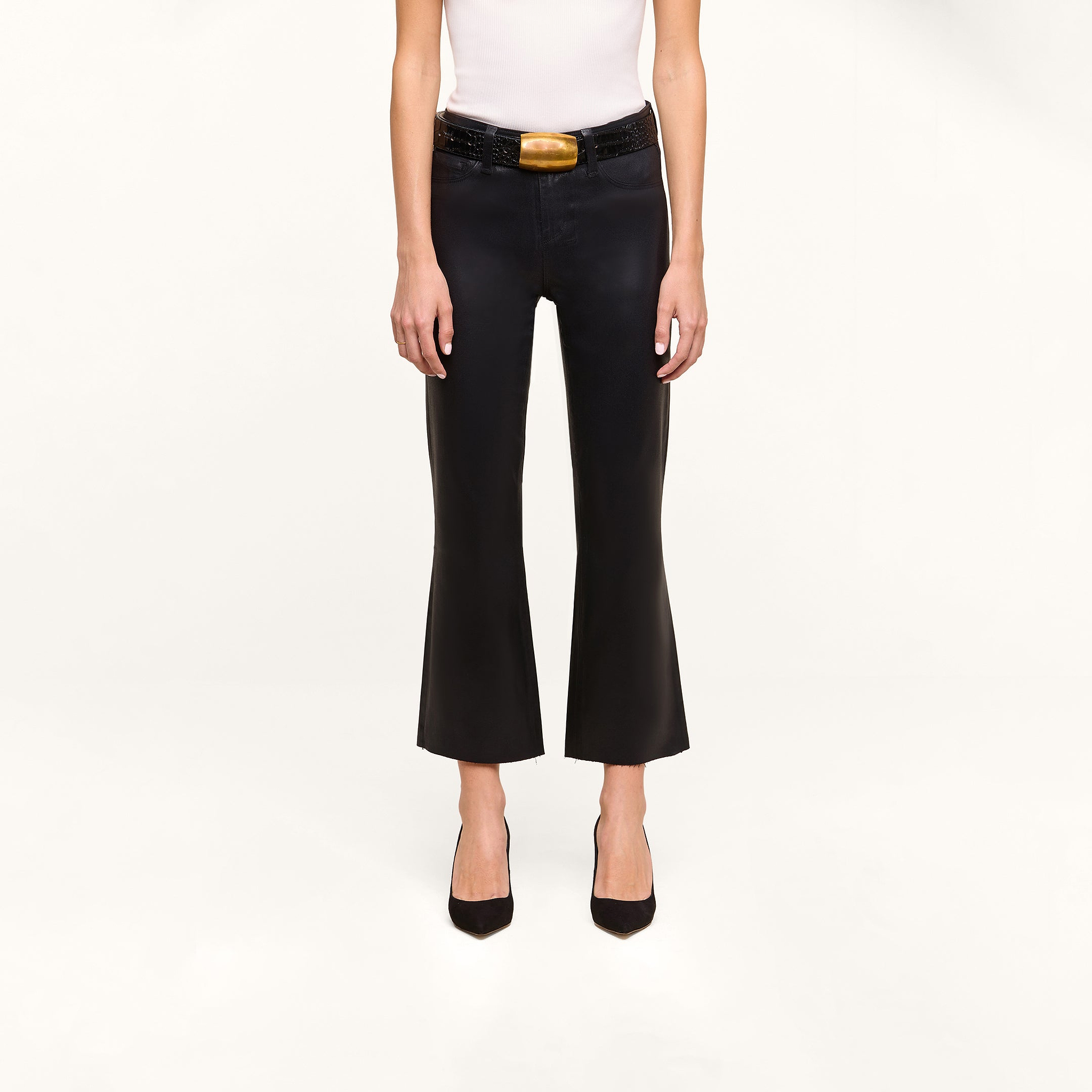 Person wearing a white sleeveless top and the Petite Kendra Coated Jean—black high-rise flared pants styled with a wide black belt featuring a gold buckle—and black pointed-toe heels, standing against a plain white background.