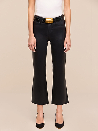 Standing against a plain beige background, a person wears a white sleeveless top, black pointed-toe shoes, a black belt with a gold buckle, and the Petite Kendra Coated Jean from the Les Petites Collection in high rise black cropped flare style.