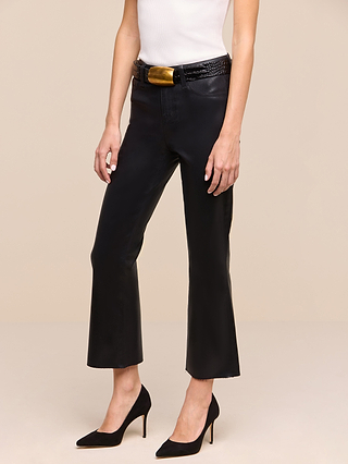 A person wearing the Petite Kendra Coated Jean from the Les Petites Collection stands against a plain beige background, styled with a white sleeveless top, a black belt with a gold buckle, and black high heels.