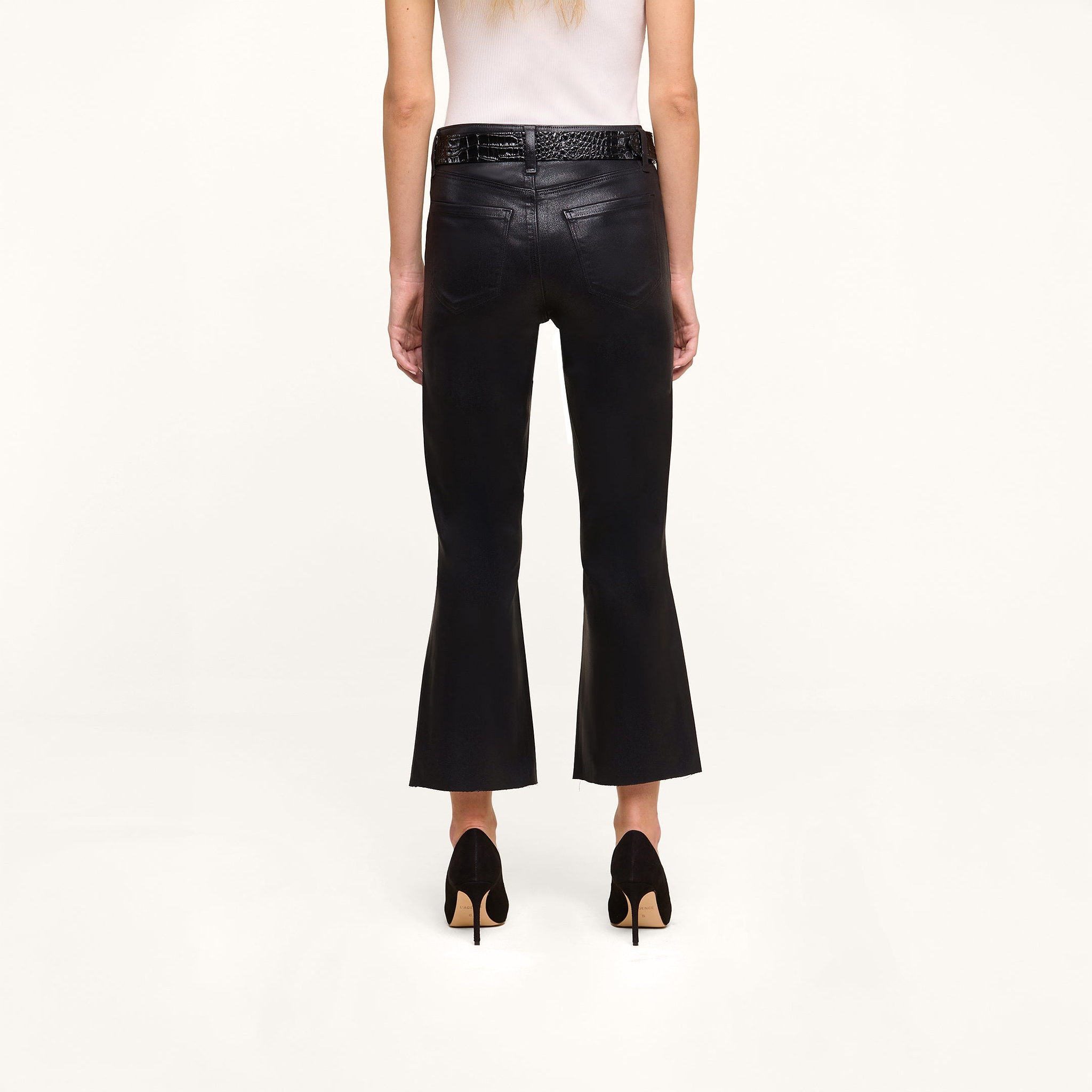 A person stands facing away against a plain white background, wearing the Petite Kendra Coated Jean in black high rise denim, paired with a white sleeveless top, black high heels, and a black braided belt.