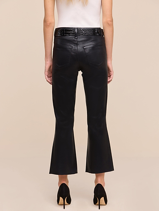 A woman wearing the Petite Kendra Coated Jean—a pair of black high rise cropped leather pants—along with a white sleeveless top, black belt, and black high-heeled shoes stands facing away against a beige background, showcasing the Les Petites Collection.