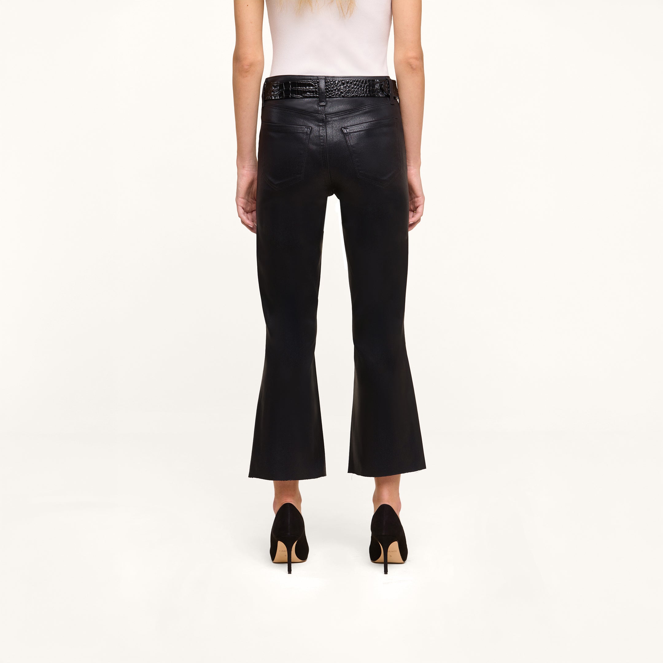 A person stands facing away against a plain white background, wearing the Petite Kendra Coated Jean in black high rise denim, paired with a white sleeveless top, black high heels, and a black braided belt.