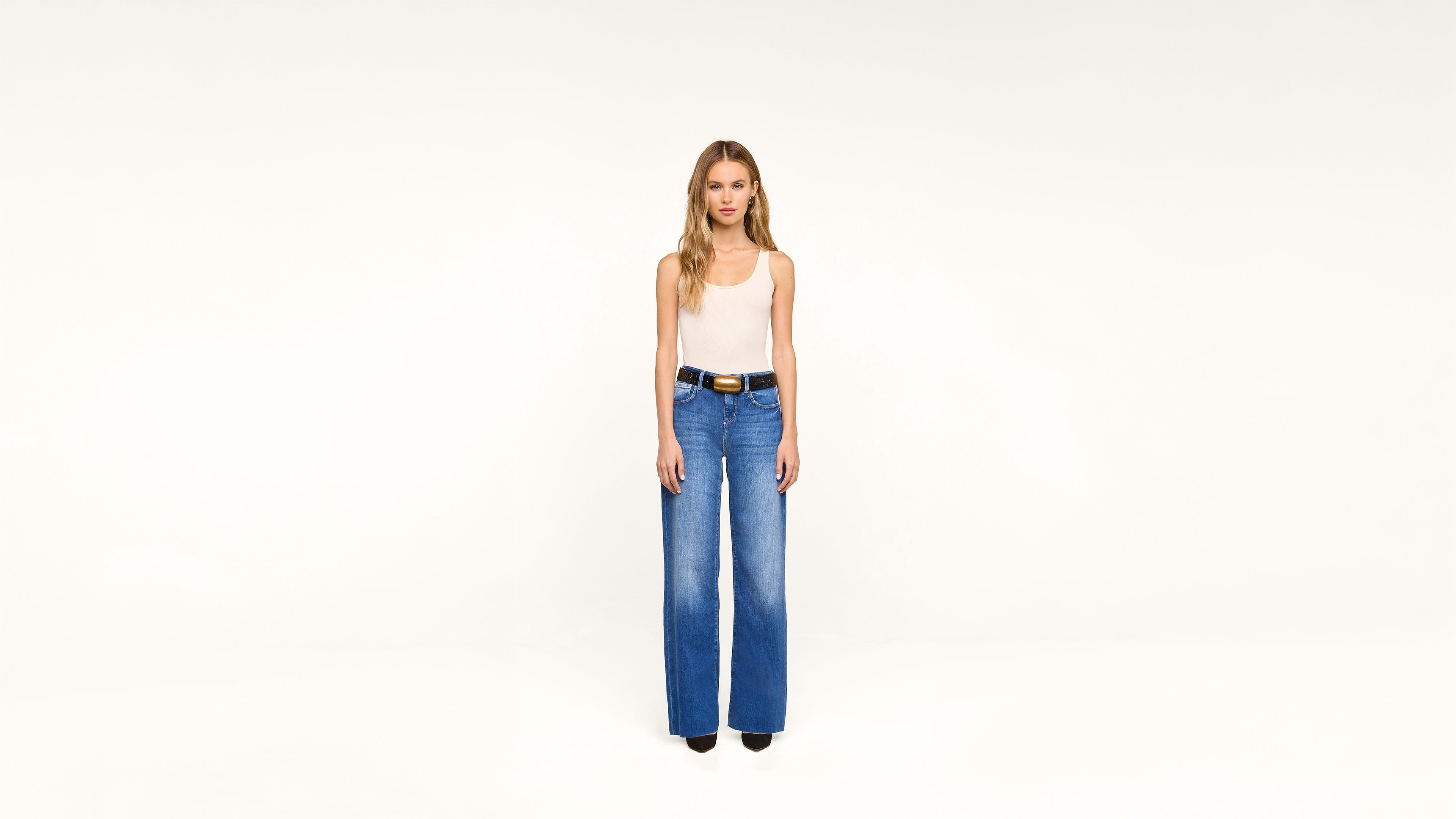 A woman stands against a plain white background, wearing a sleeveless beige top, high-waisted blue wide-leg jeans, and black shoes. She has long, wavy blonde hair and a neutral expression.