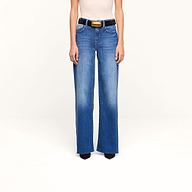 A person is wearing the Petite Scottie Wide-Leg Jean in blue, styled with a white sleeveless top, a black belt featuring a large gold buckle, and black pointed shoes, standing against a plain white background.