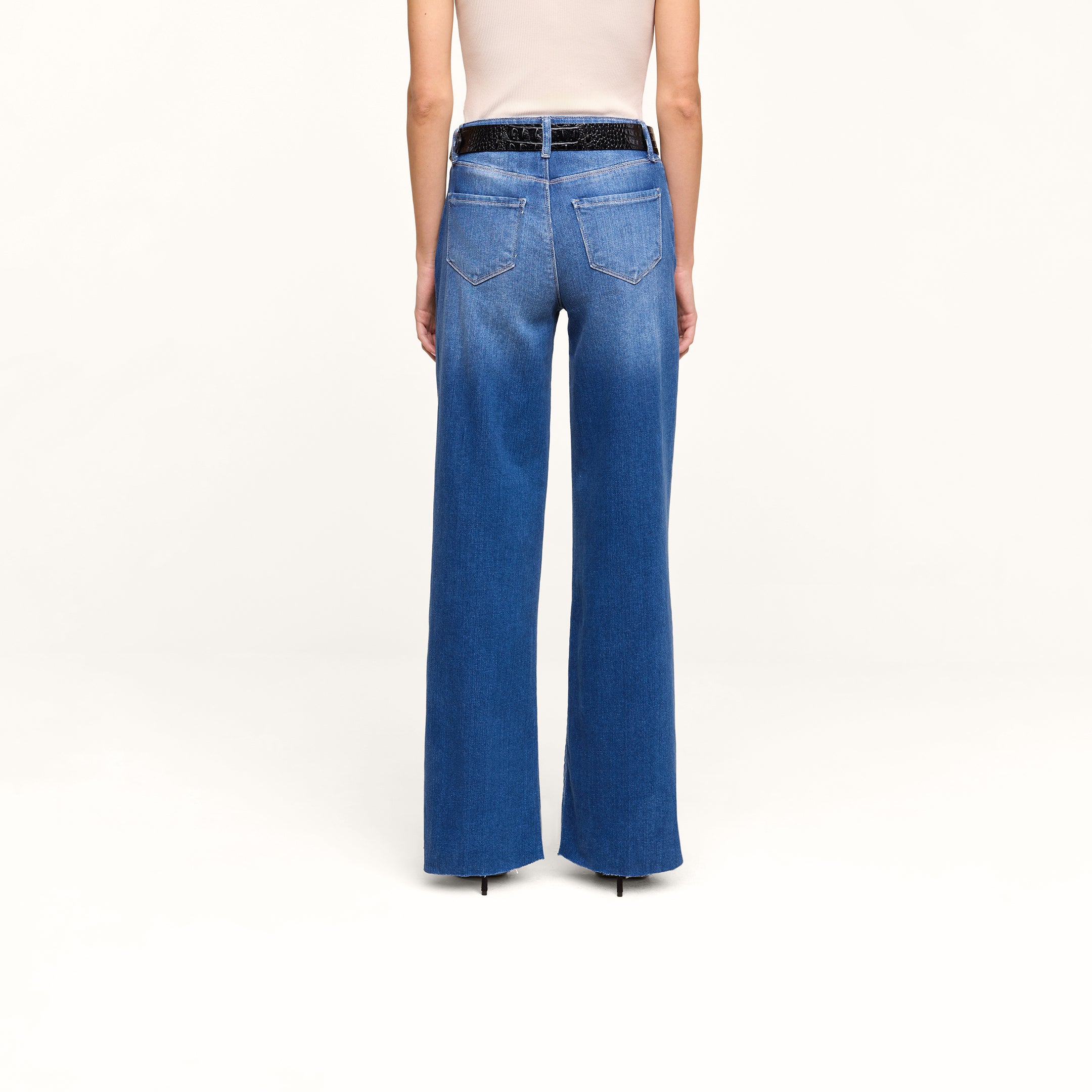 Wearing the Petite Scottie Wide-Leg Jean in blue stretch denim and a white sleeveless top, this person is shown from the back. The high-rise jeans feature two back pockets and a black belt. The background is plain white.