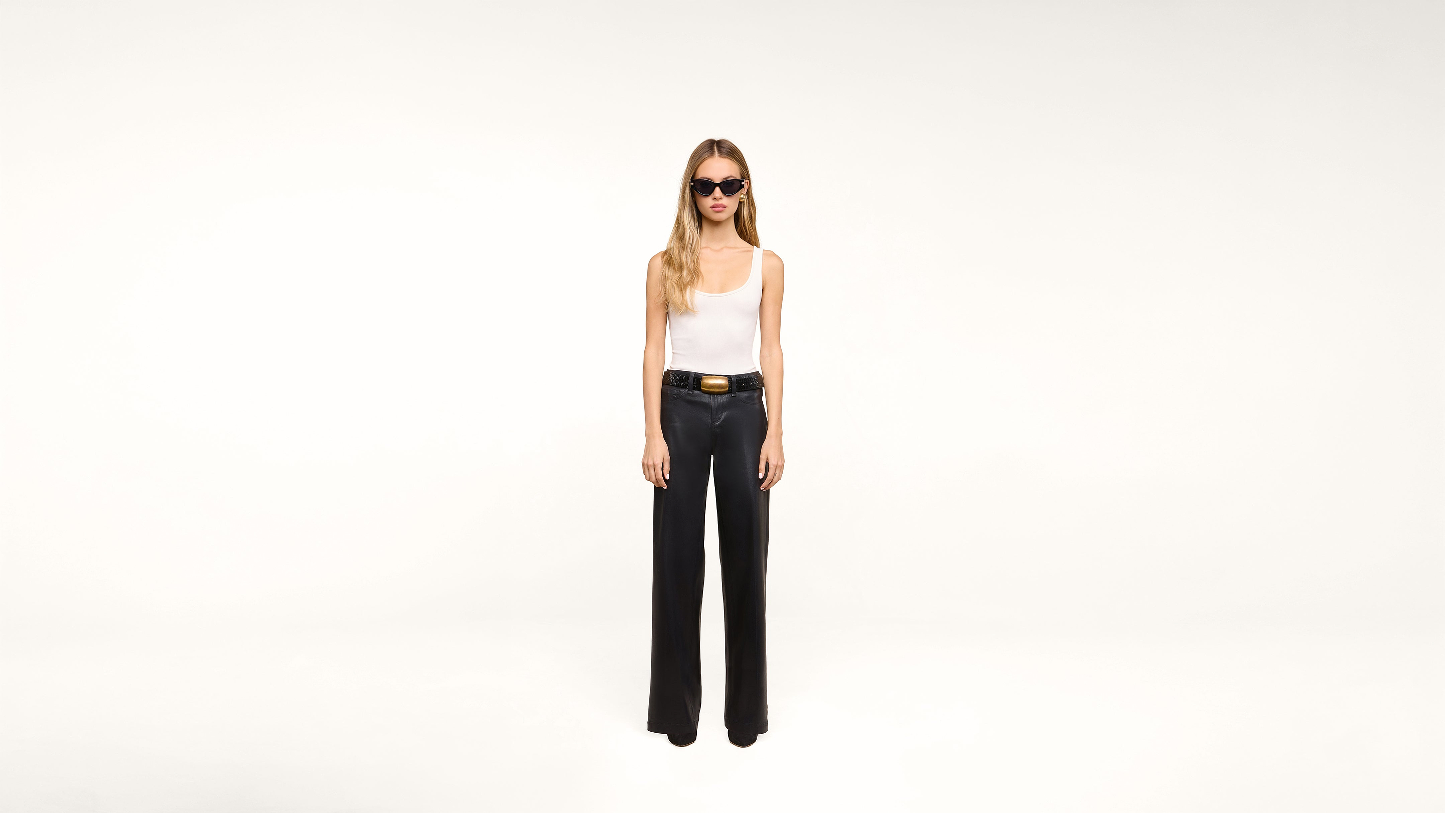 A woman wearing sunglasses, a white tank top, black wide-leg pants, and a black belt stands against a plain white background.