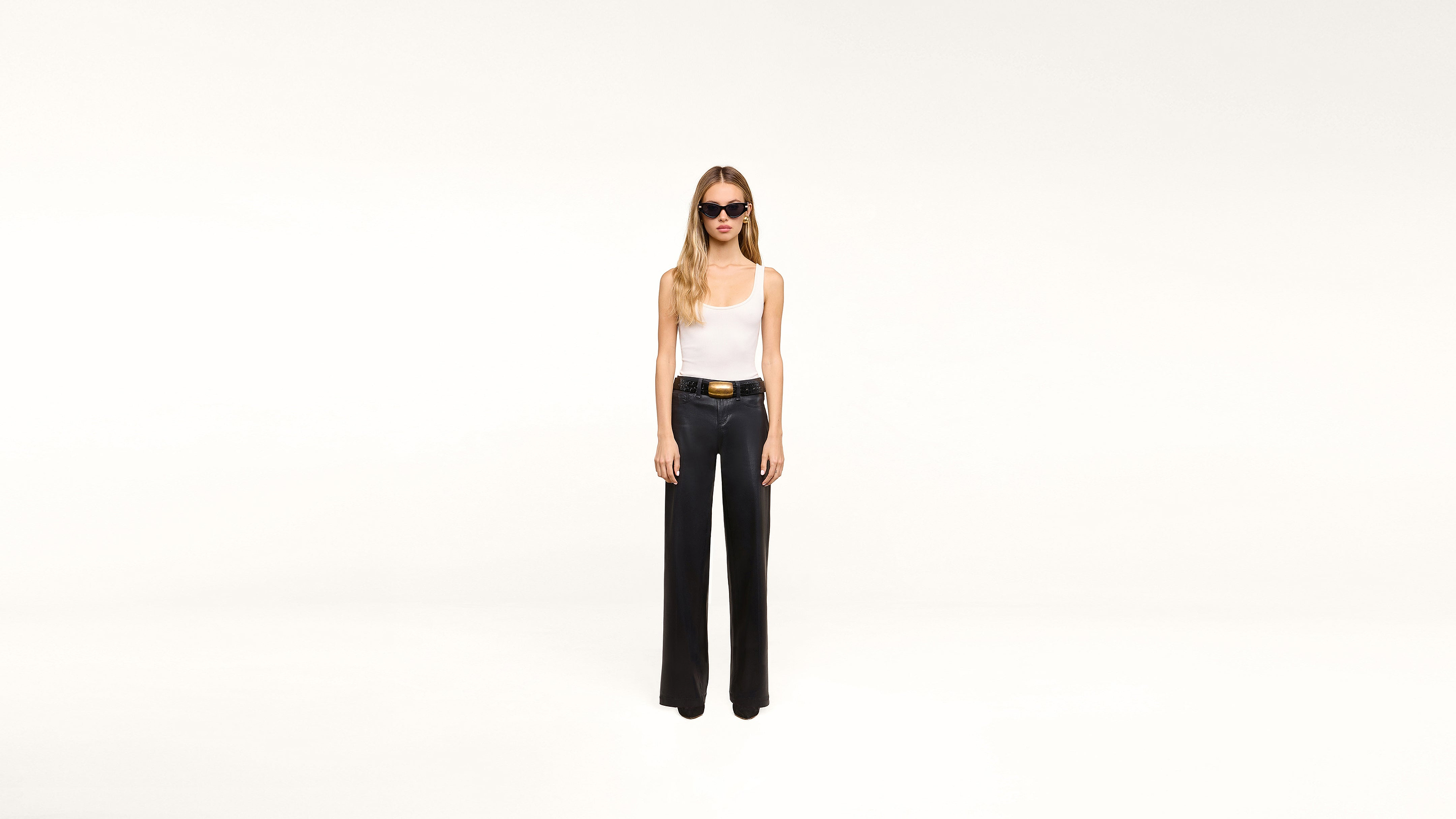 A woman wearing sunglasses, a white tank top, and black wide-leg pants paired with a stylish black belt stands confidently against a plain white background.