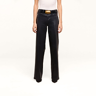 A person is wearing the Petite Scottie Coated Jean, styled in a high rise black wide-leg silhouette, paired with a white sleeveless top and accessorized with a black belt featuring a large gold buckle, standing against a plain white background.