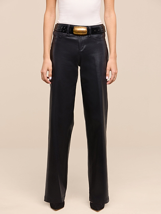A person wearing the Petite Scottie Coated Jean from the Les Petites Collection—high-rise black wide-leg pants—styled with a white sleeveless top and a black belt featuring a large rectangular gold buckle, standing against a beige background.