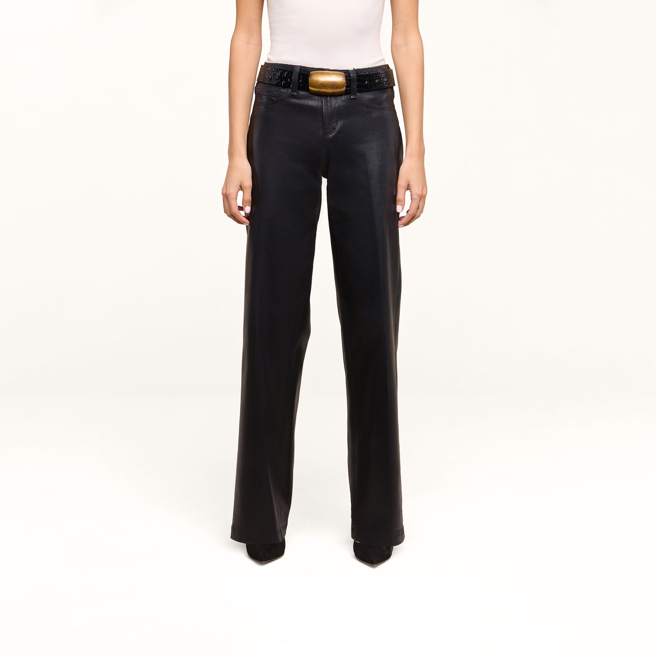 A person is wearing the Petite Scottie Coated Jean, styled in a high rise black wide-leg silhouette, paired with a white sleeveless top and accessorized with a black belt featuring a large gold buckle, standing against a plain white background.