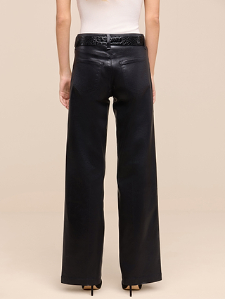 From the Les Petites Collection, a person is shown from the back, standing against a plain beige background, wearing the Petite Scottie Coated Jean—a high-waisted, black wide-leg style in coated denim for petites—paired with a black textured belt and a white sleeveless top.