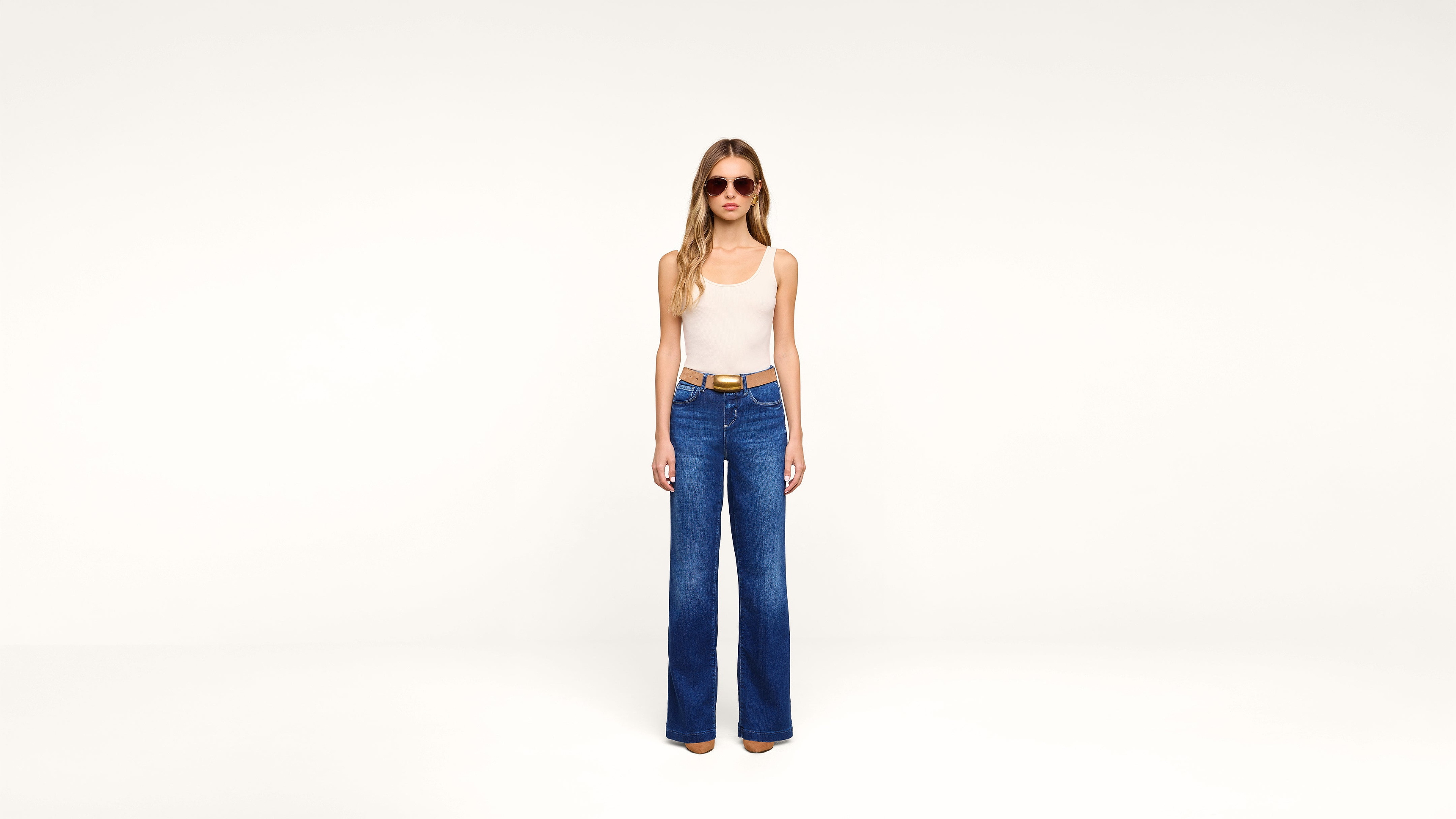 A woman wearing sunglasses, a white sleeveless top, blue high-waisted wide-leg jeans, and tan shoes stands against a plain white background.