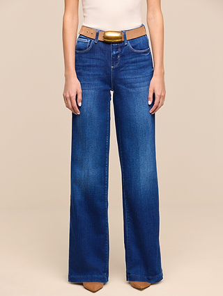 A person wearing the Petite Scottie Wide-Leg Jean from the Les Petites Collection in blue, paired with a beige belt featuring a gold buckle and tan pointed-toe shoes, stands against a plain beige background.