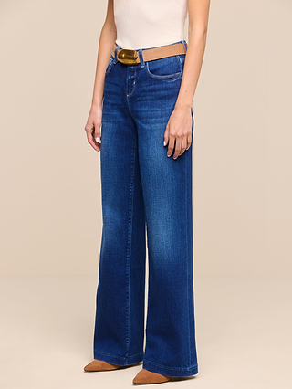 A person wearing the Petite Scottie Wide-Leg Jean from the Les Petites Collection in blue, styled with a brown belt, a sleeveless white top, and brown pointed-toe shoes, stands against a plain beige background.
