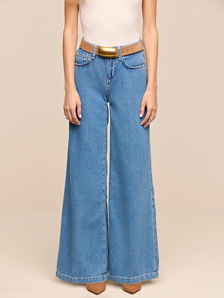 A woman wearing the Petite Quincy Wide-Leg Jean from the Les Petites Collection in denim blue, a beige sleeveless top, a tan belt, and pointed-toe brown shoes stands against a plain beige background.