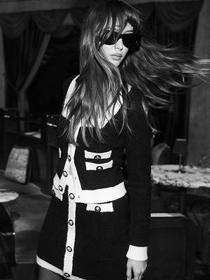 A woman with long hair and sunglasses poses indoors, wearing the L'AGENCE Lottie Knit Mini Skirt in a black-and-white photo, her hair flowing as she turns her head.