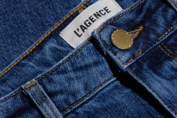 Close-up of blue denim jeans with a metal button and a white fabric label reading LAGENCE sewn near the waistband. The jeans have visible orange stitching and a classic five-pocket design.
