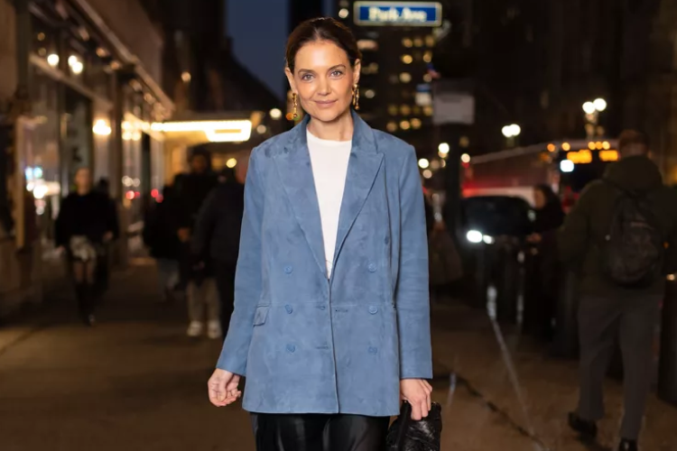 A woman in a blue blazer and white shirt stands on a city sidewalk at night, smiling. People, streetlights, and cars are visible in the blurred background.