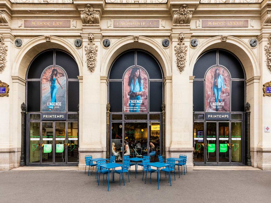 Outdoor view of a grand building with three large arched windows, posters of a woman in blue outfits above each entrance, and blue tables and chairs arranged on the sidewalk in front.