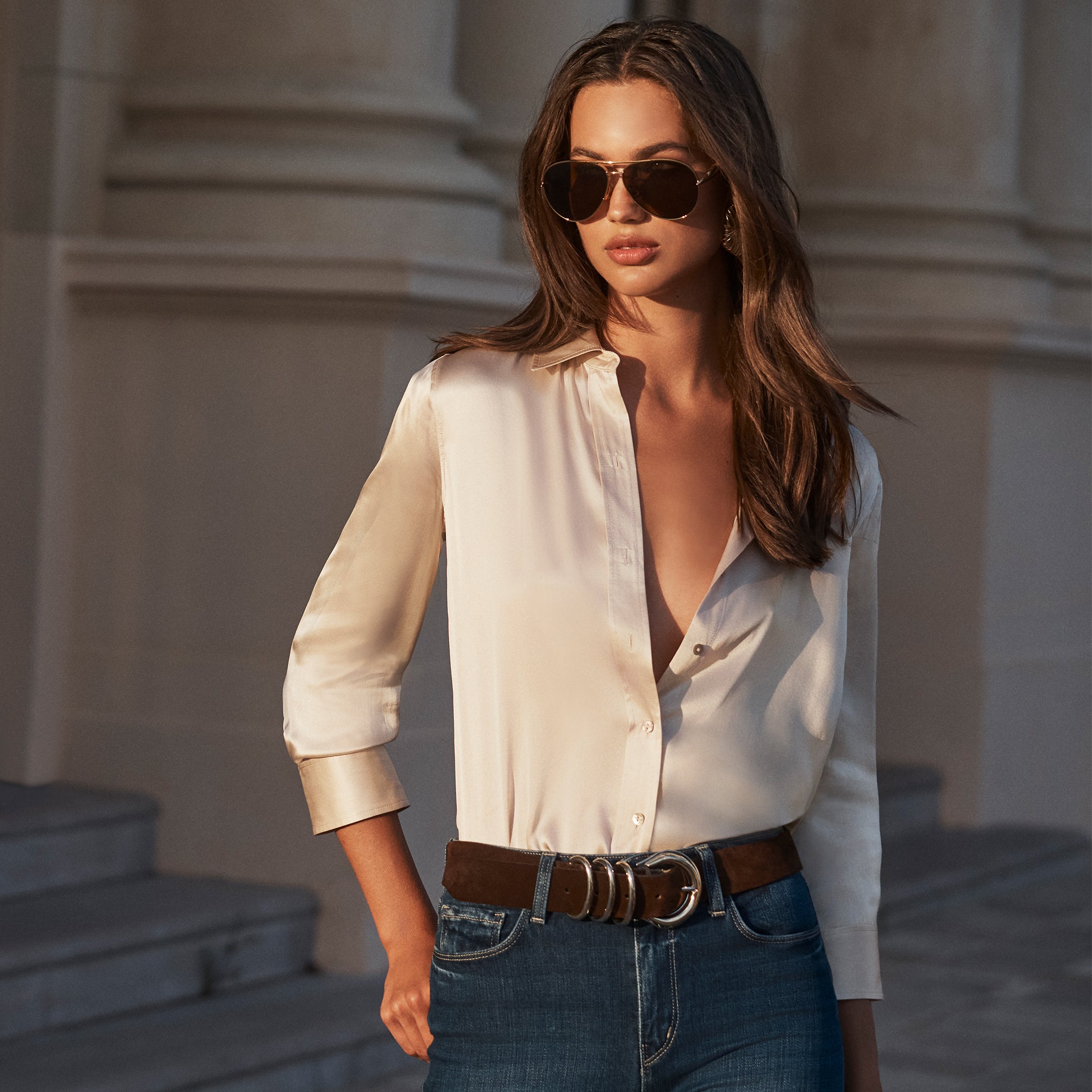 A person wearing a long-sleeved, champagne-colored Dani Silk Blouse tucked into blue jeans with a brown leather belt and aviator sunglasses.