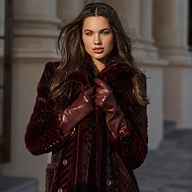 A woman with long brown hair stands confidently outside in front of a stone building, wearing a dark maroon fur coat and the luxurious Carmen Gloves, looking directly at the camera.