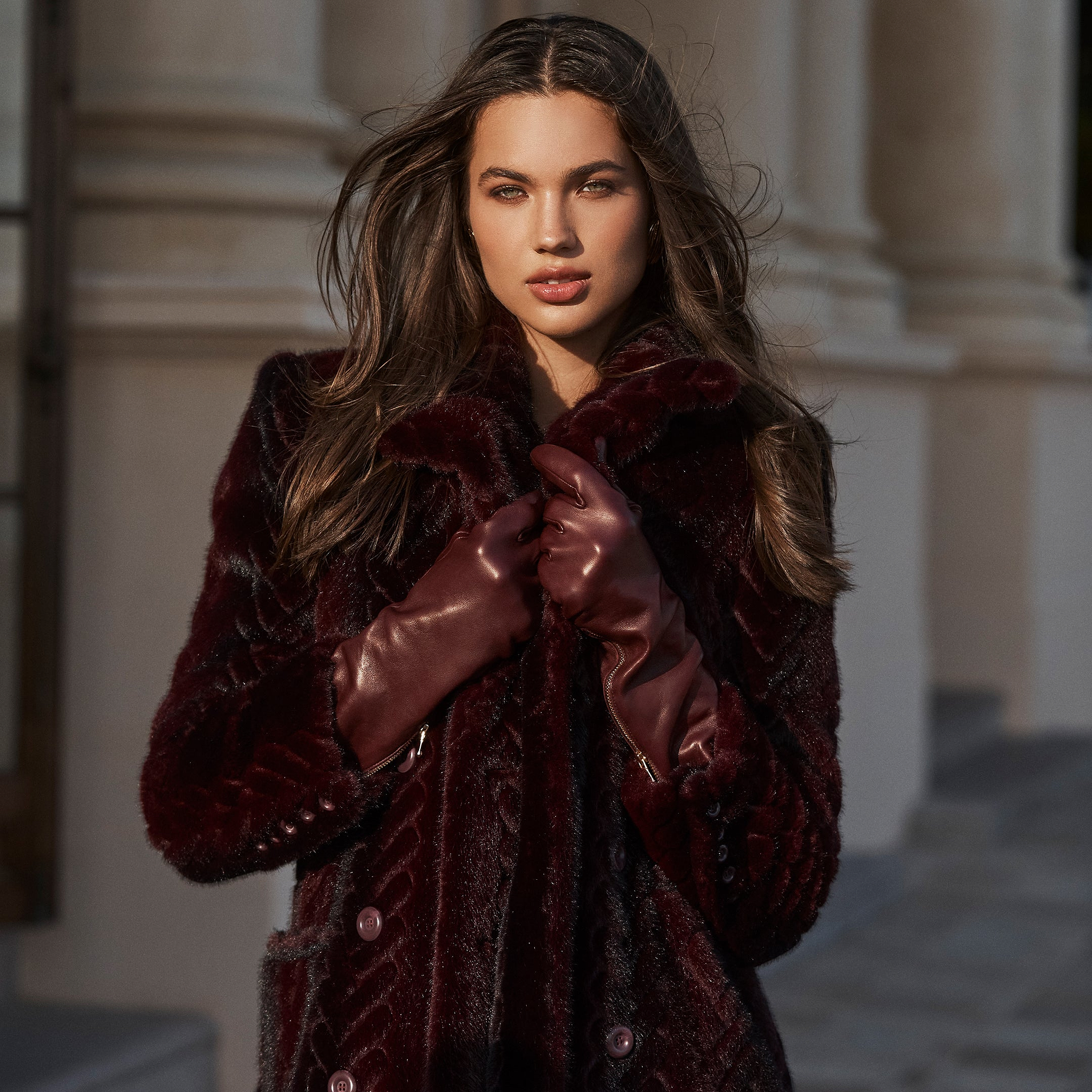 A woman with long brown hair stands confidently outside in front of a stone building, wearing a dark maroon fur coat and the luxurious Carmen Gloves, looking directly at the camera.