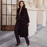 A person wearing a long, deep purple Icelyn Faux Fur Coat with a textured pattern, dark trousers, and gloves, standing on stone steps as part of the Fall 2026 collection.