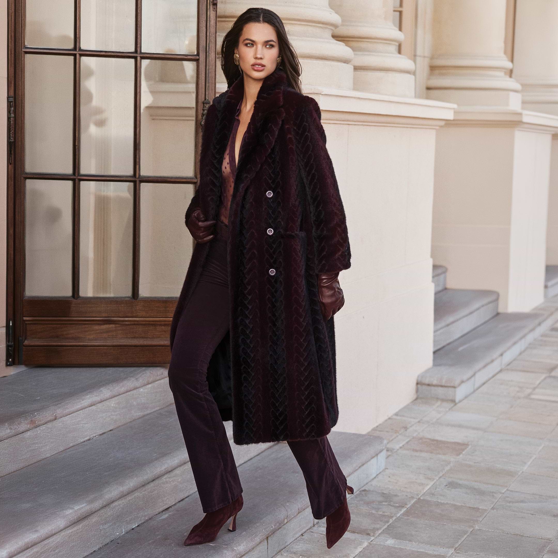A person wearing a long, deep purple Icelyn Faux Fur Coat with a textured pattern, dark trousers, and gloves, standing on stone steps as part of the Fall 2026 collection.