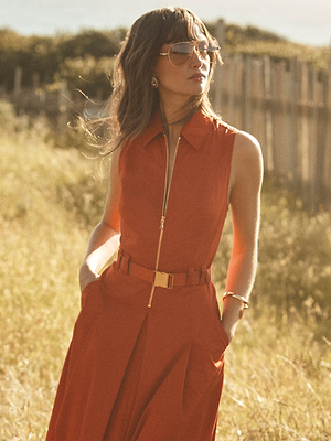 A woman stands outdoors in sunlight wearing the Kolton Dress—a sleeveless, collared burnt orange midi dress with pockets and a belt—paired with aviator sunglasses and earrings, against a blurred wooden fence and grassy background.