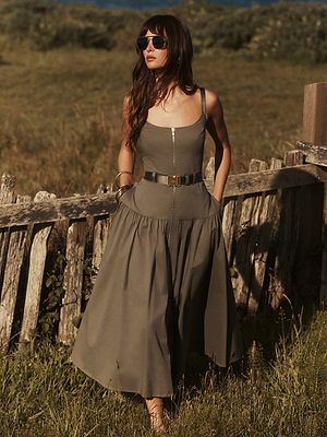 A woman in the Rima Dress—an olive green stretch poplin maxi with a belt and sunglasses—stands by a rustic wooden fence in a grassy outdoor setting.