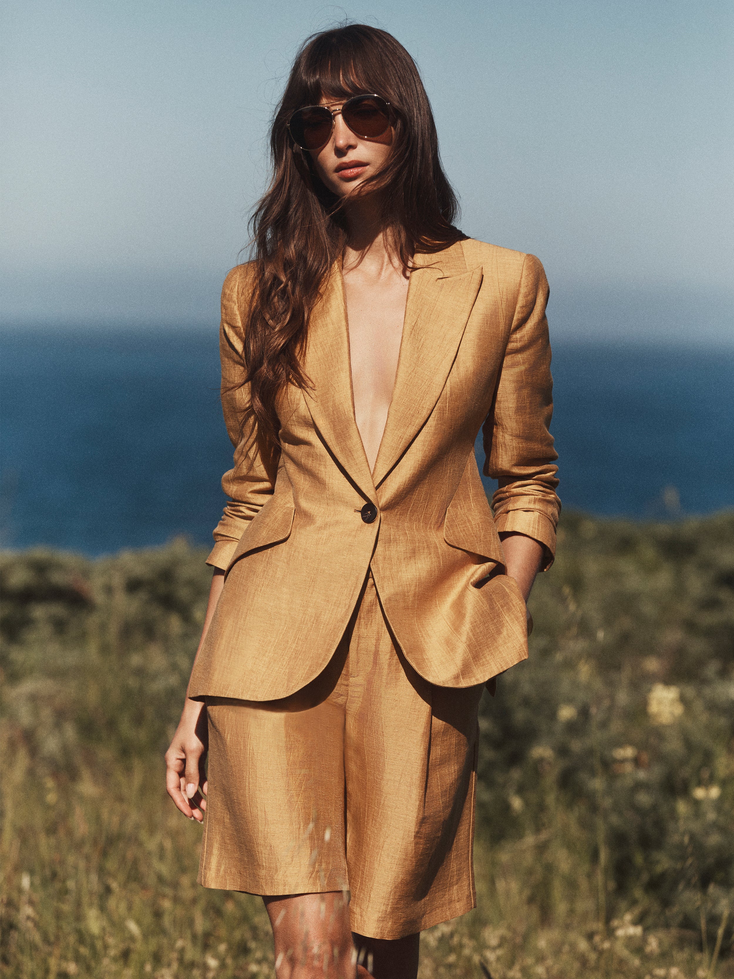 Una donna posa con disinvoltura allaperto, sullo sfondo del verde e delloceano, indossando occhiali da sole e il blazer dorato Chamberlain in misto lino, abbinato a pantaloncini al ginocchio dello stesso colore.
