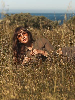 A woman with long brown hair and sunglasses lies in tall grass, wearing the olive green Carine Top. Trees, blue sky, and water appear in the background.