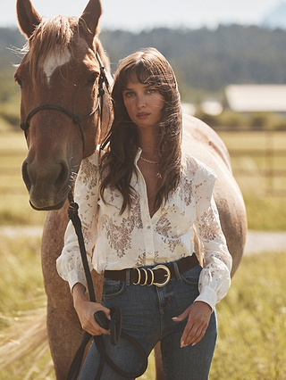 A woman wearing the Jenica Lace Blouse and jeans stands outdoors holding the reins of a brown horse in a sunlit field, with blurred trees and a building in the background.