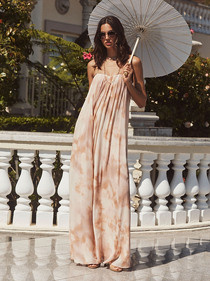 Une femme portant des lunettes de soleil et une robe fluide sans bretelles Raine avec corset intégré se tient à lextérieur en tenant un parasol blanc, devant une balustrade blanche avec de la verdure luxuriante et des fleurs derrière elle.