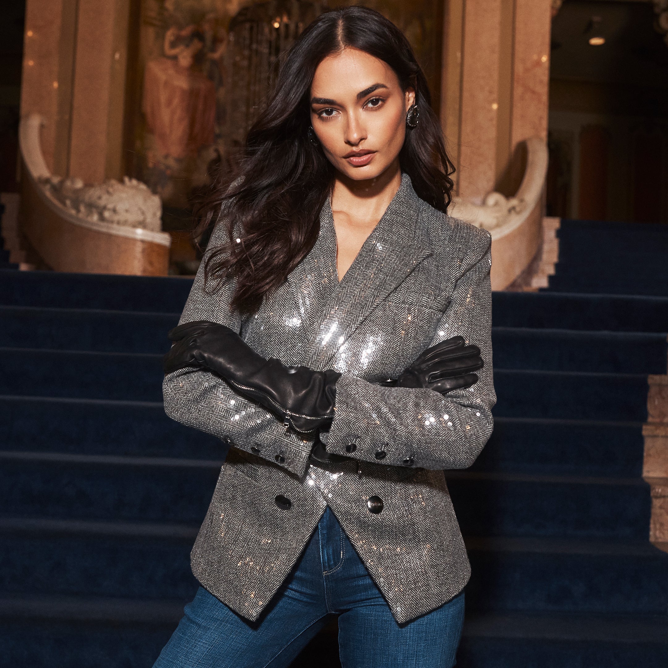 A person wears a silver Kyomi Sequined Blazer and black leather gloves while standing with arms crossed in front of a blue carpeted staircase.