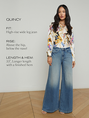 A woman stands indoors wearing high-rise, wide-leg blue jeans and a colorful patterned blouse. Text on the image describes the jeans’ fit, rise, and length details. She stands on a wooden floor against a plain wall.