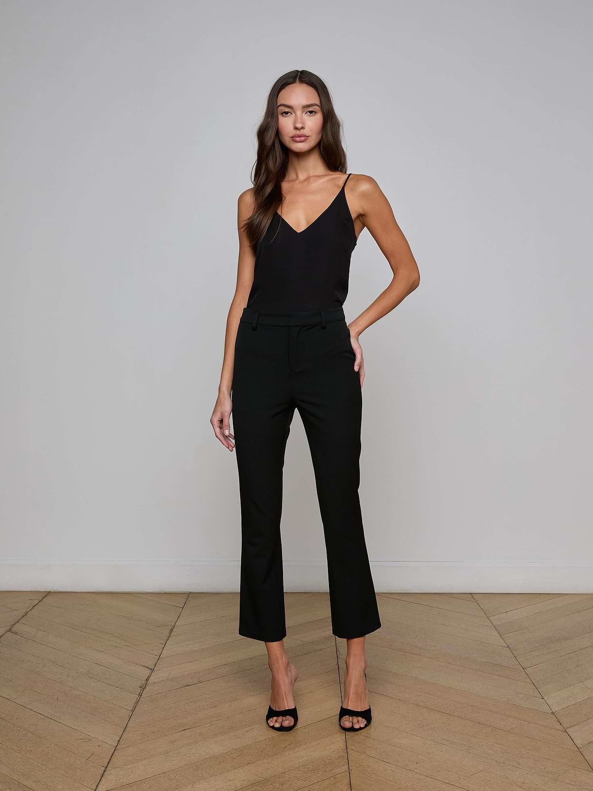 A woman with long hair wears a black sleeveless top and the Ketra Bootcut Trouser in black, crafted from midweight suiting fabric, paired with black high-heeled sandals as she stands against a plain white background.