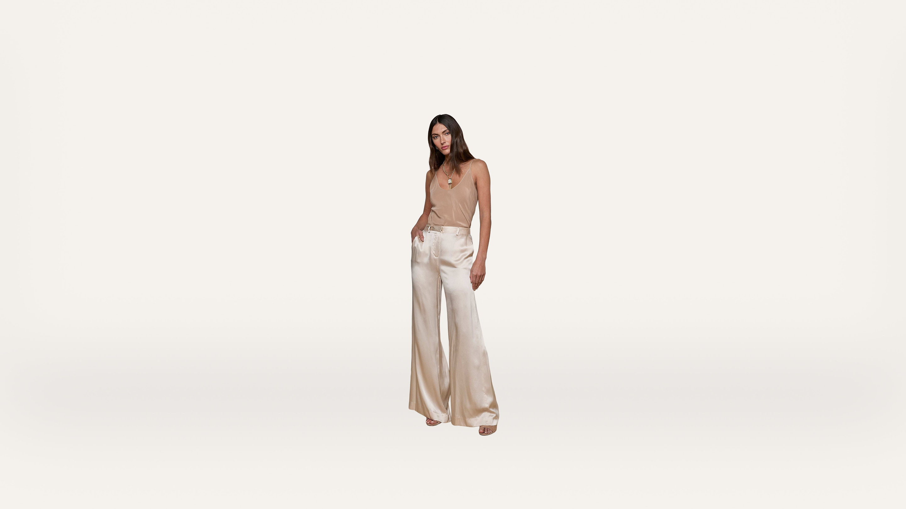Woman standing against a plain white background, wearing a beige sleeveless top and high-waisted, wide-leg cream satin pants, with one hand in her pocket and neutral facial expression.
