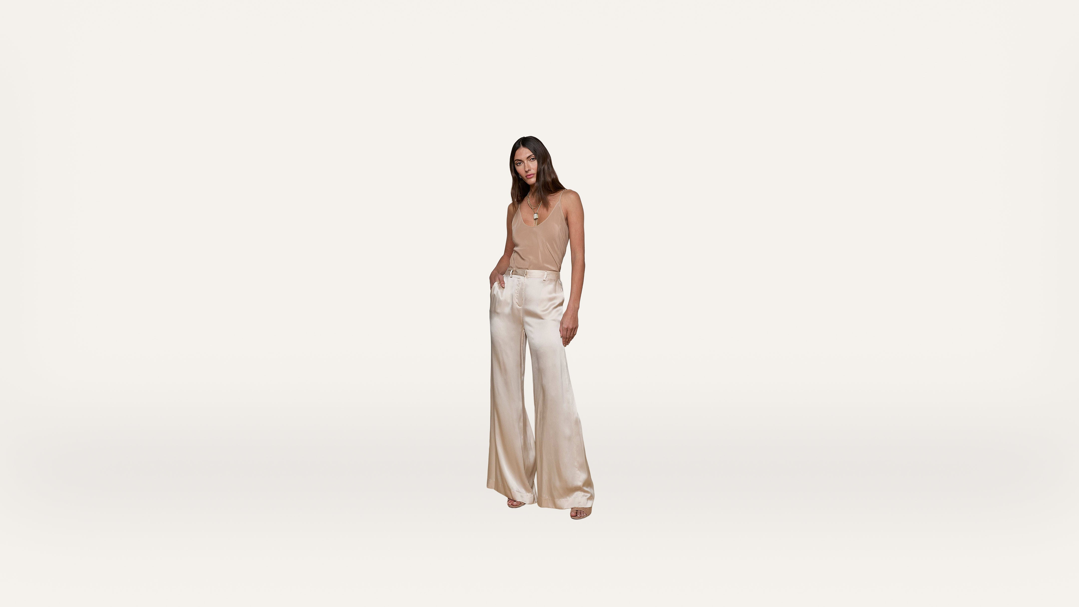 Woman standing against a plain white background, wearing a beige sleeveless top and high-waisted, wide-leg cream satin pants, with one hand in her pocket and neutral facial expression.