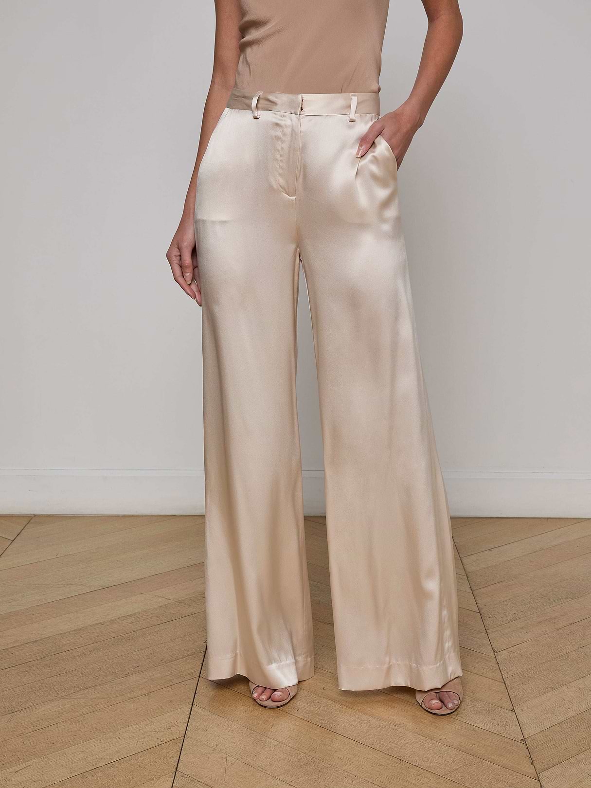 A woman stands against a plain white background, one hand in her pocket, wearing the Gavin Silk Wide-Leg Pant in a light champagne hue paired with a sleeveless top.