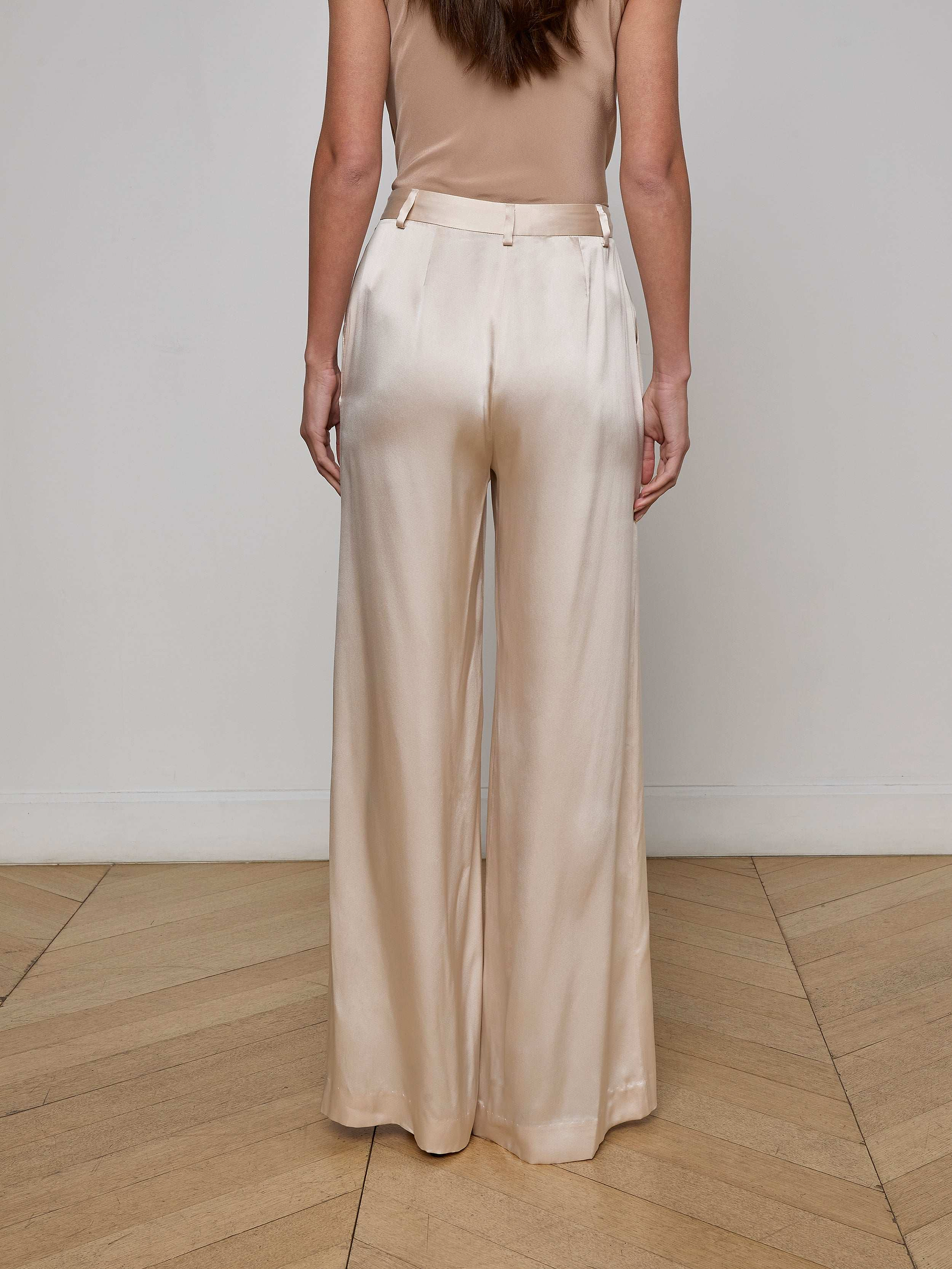 A woman stands facing away, wearing the Gavin Silk Wide-Leg Pant in a champagne color paired with a sleeveless beige top, set against a plain white background.