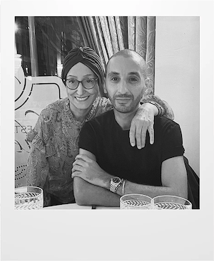 A woman wearing glasses and a headscarf smiles with her arm around a man in a black t-shirt. They are seated at a table with patterned glasses, in a warmly lit indoor setting with curtains behind them.