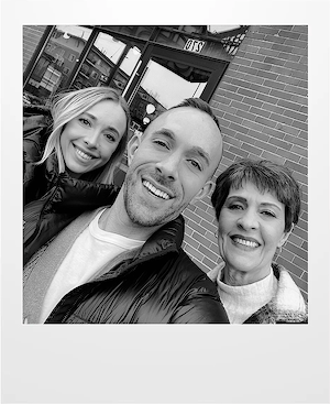 Three people smile for a selfie outside a brick building. The two women and one man, all dressed warmly, are closely grouped and appear happy. The photo is in black and white.