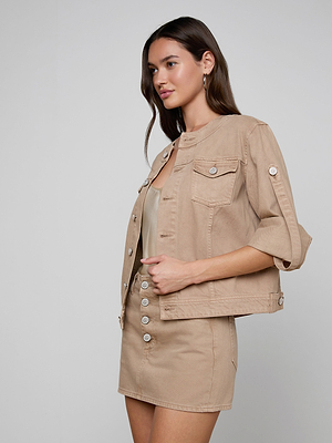 A woman with long brown hair wears the Anouk Jean Jacket with roll-tab sleeves over a beige button-front mini skirt, standing against a plain light gray background.