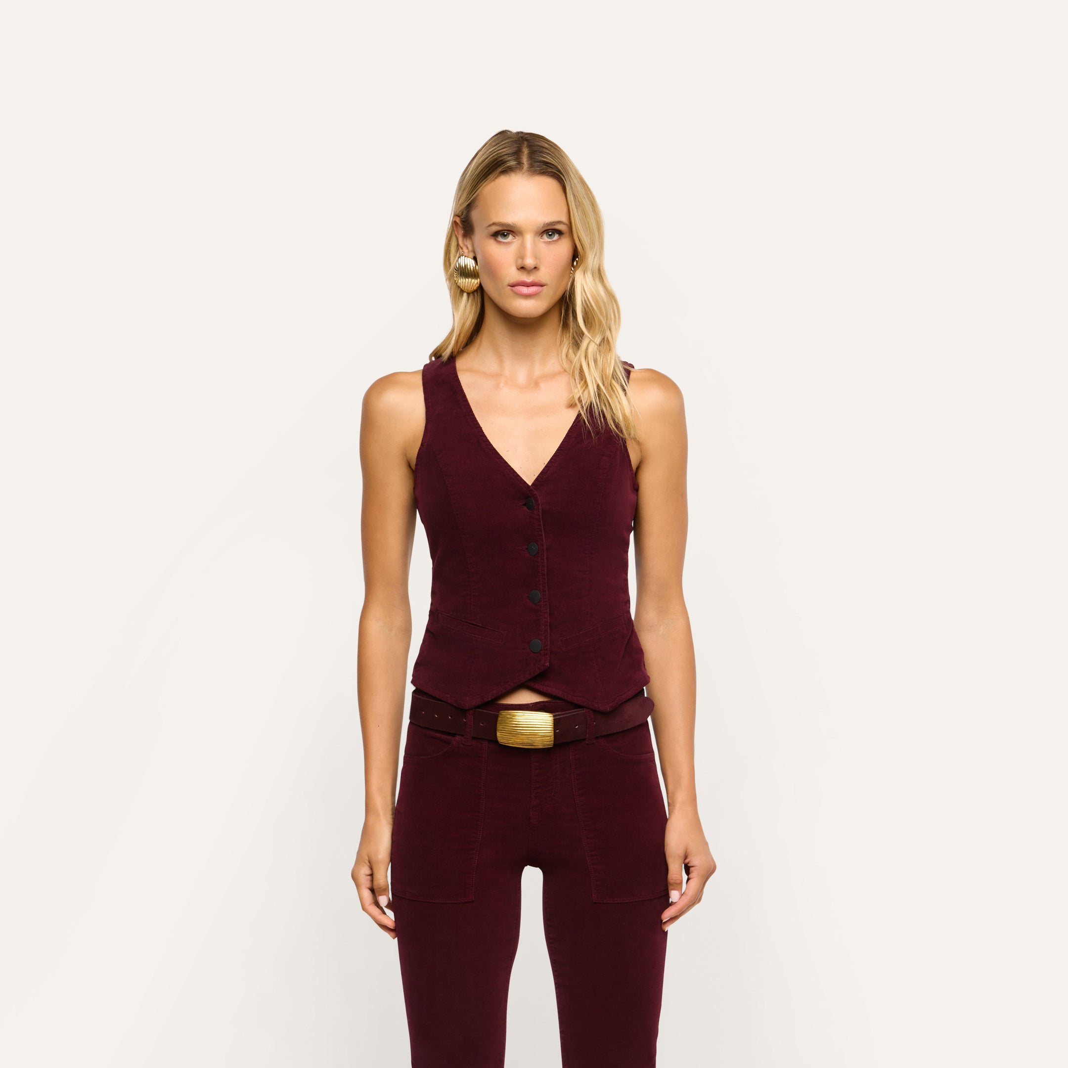 A woman wears a burgundy stretch corduroy Yelena Corduroy Vest and matching trousers with a gold-buckled belt.