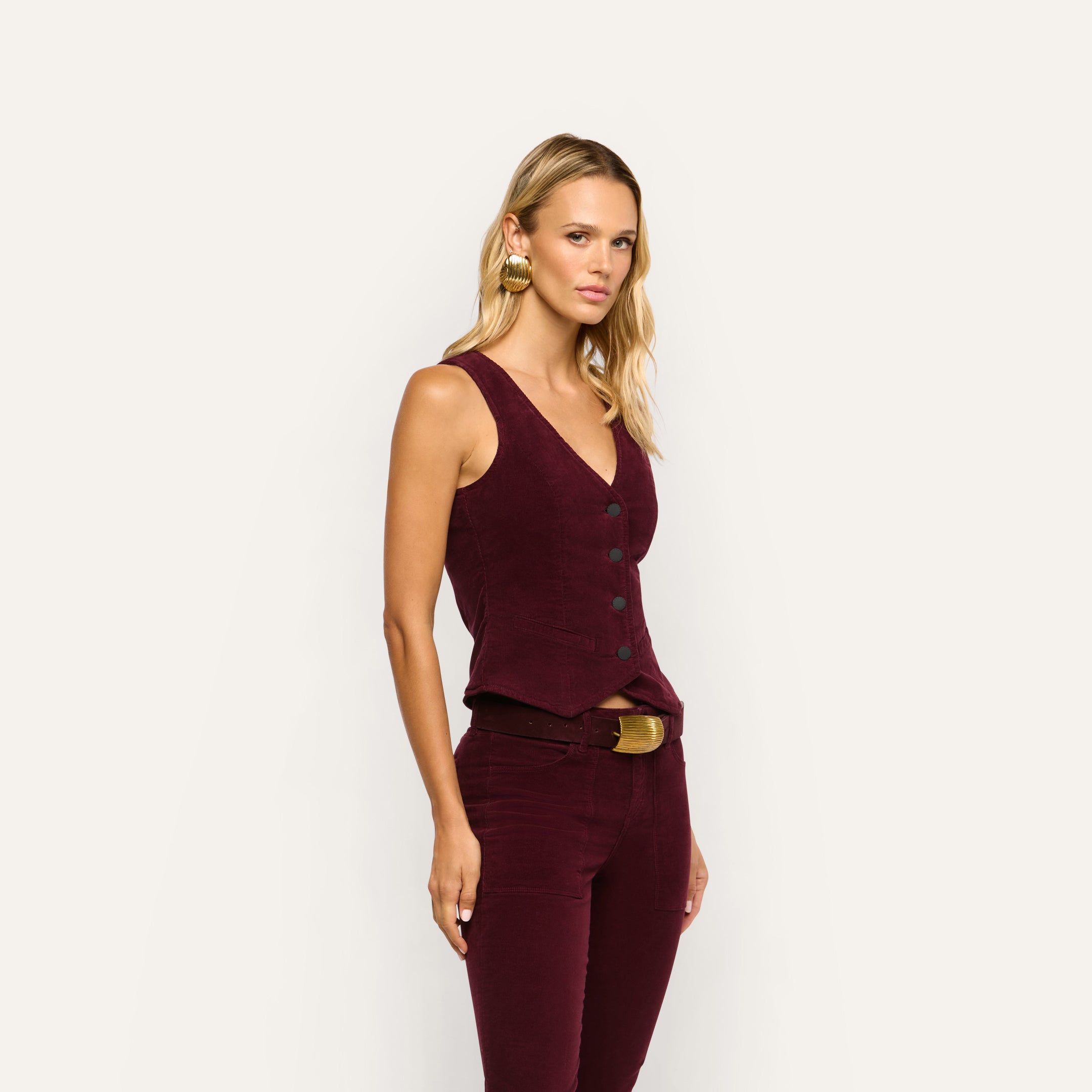 A person wears a tailored dark red stretch corduroy Yelena Corduroy Vest and matching pants with a gold-buckle belt.