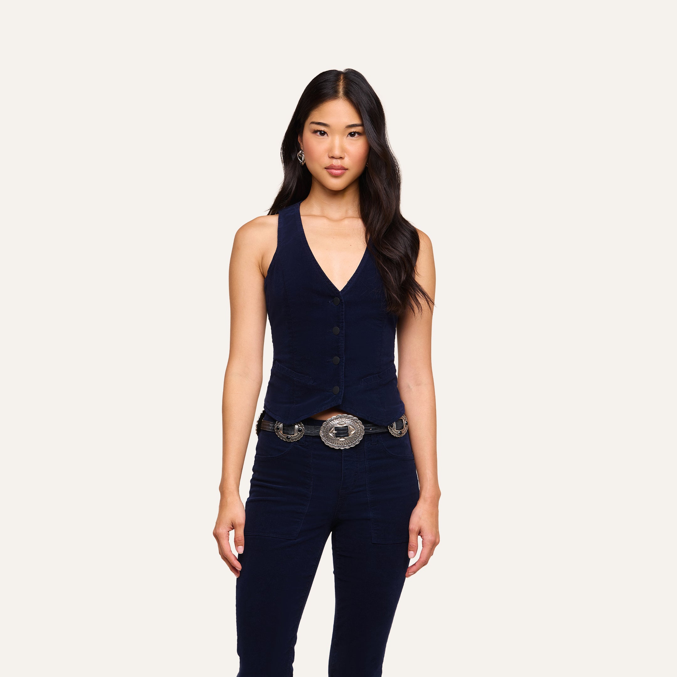 A woman with long dark hair wears the Yelena Vest, a Western-inspired sleeveless style in navy blue, paired with matching pants and a decorative belt featuring large silver buckles, standing against a plain light background.